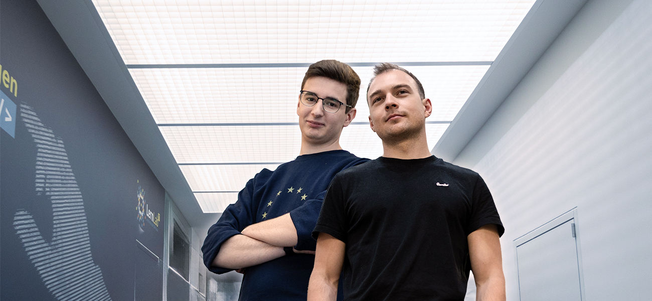 Zwei IT-Studenten stehen in einem Flur mit Rasterdecke im Bürogebäude der DRV Bund. Einer trägt ein schwarzes Shirt, der andere ein dunkelblaues Sweatshirt mit gelben Sternen.