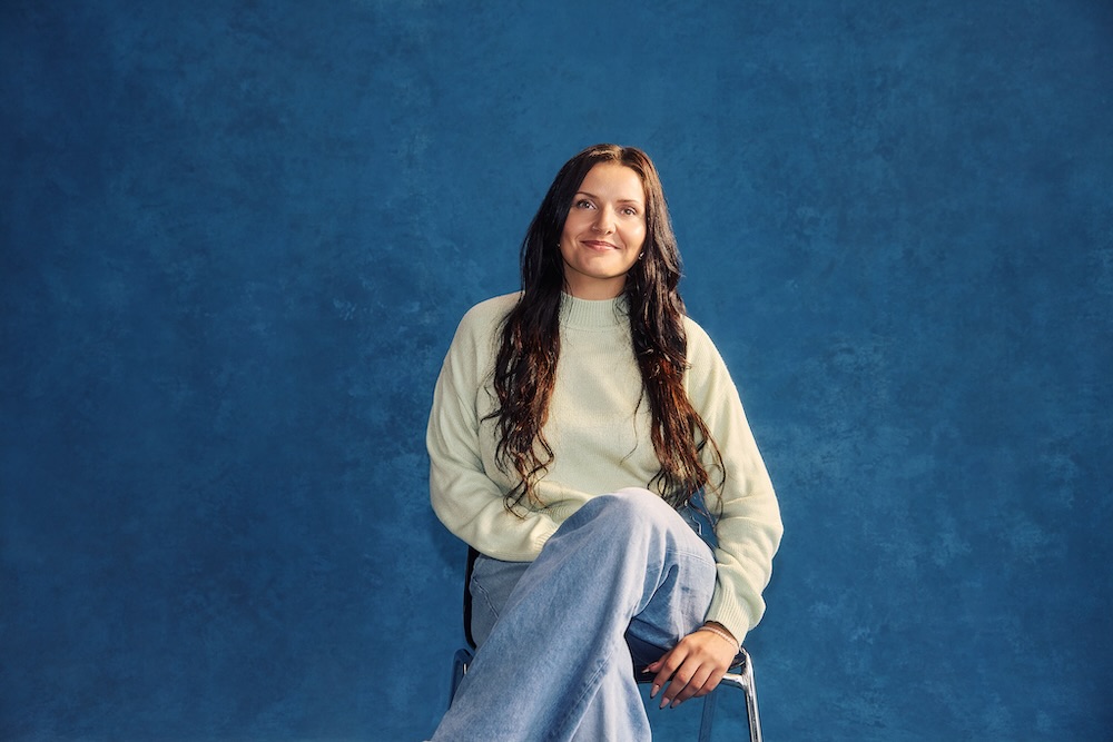 Studentin mit langem offenen Haar sitzt lächelnd auf einem Hocker vor blauem Hintergrund. Sie trägt einen beigen Pullover und eine blaue Jeans.