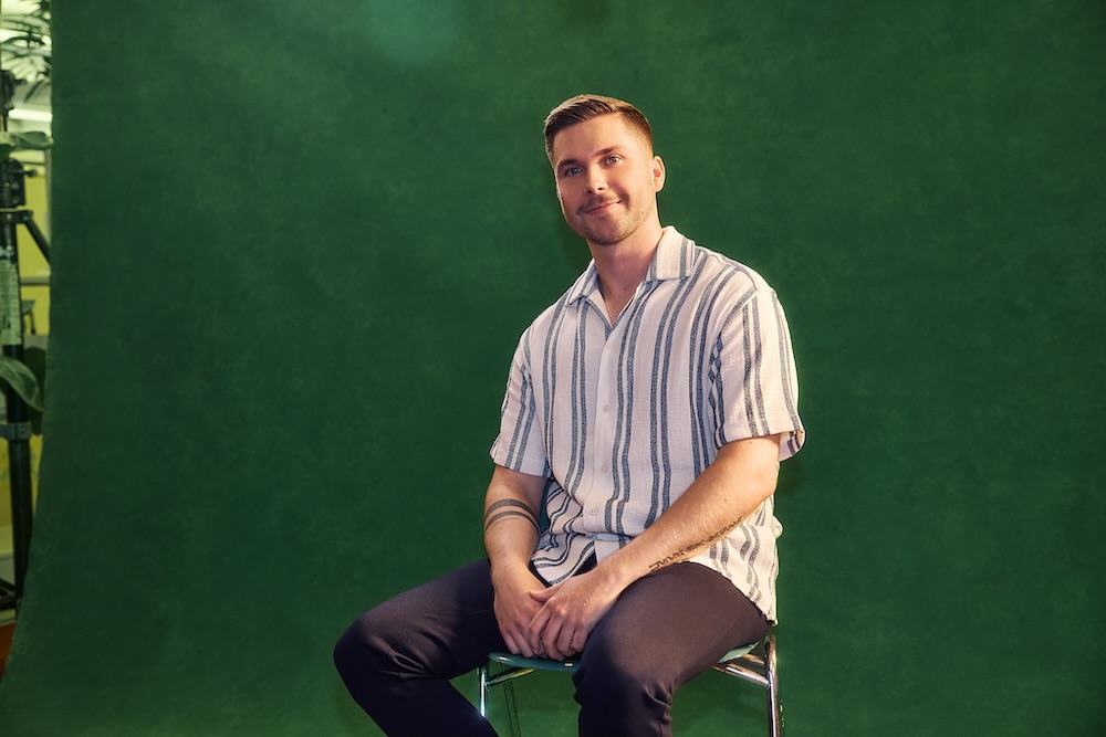 Student sitzt lächelnd auf einem Hocker vor einem grünen Hintergrund. Er trägt ein blau-weiß gestreiftes kurzärmliges Hemd und eine schwarze Hose.