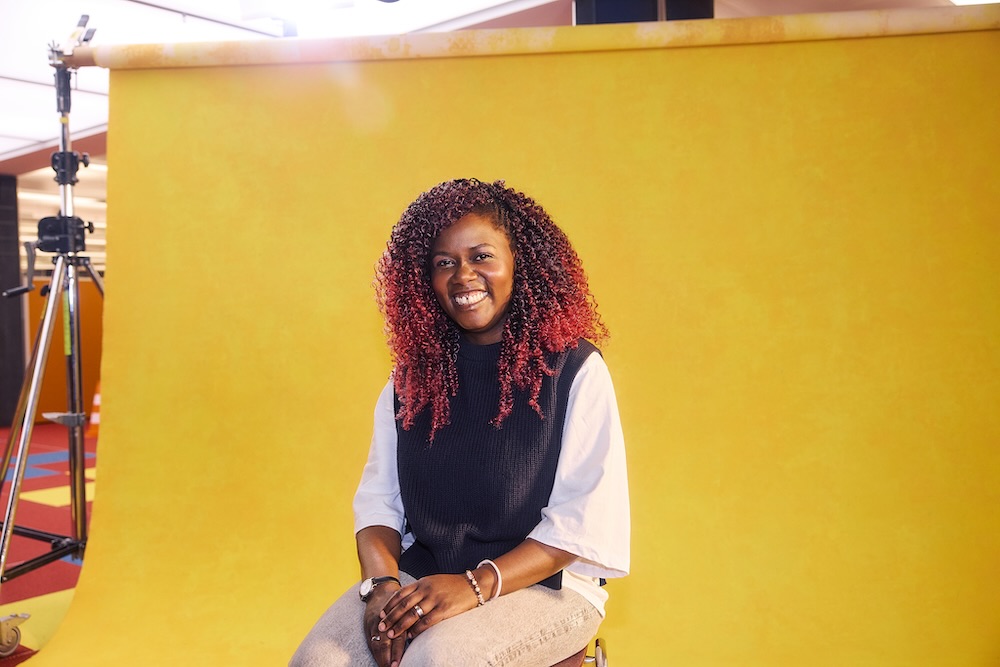 Auszubildende sitzt lächelnd auf einem Hocker vor gelbem Hintergrund in einem Fotostudio. Sie trägt einen schwarzen Strickpullunder und darunter ein weißes T-Shirt.