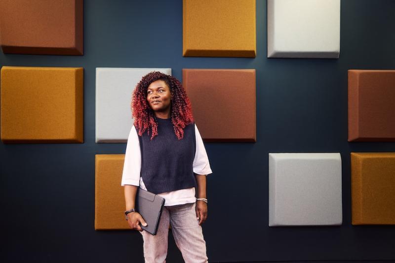 Die Auszubildende Sumaya steht im DRV Bund Gebäude in Berlin mit einem Laptop in der Hand. Sie steht vor einer Wand mit bunten Quadraten auf dunklem Hintergrund.