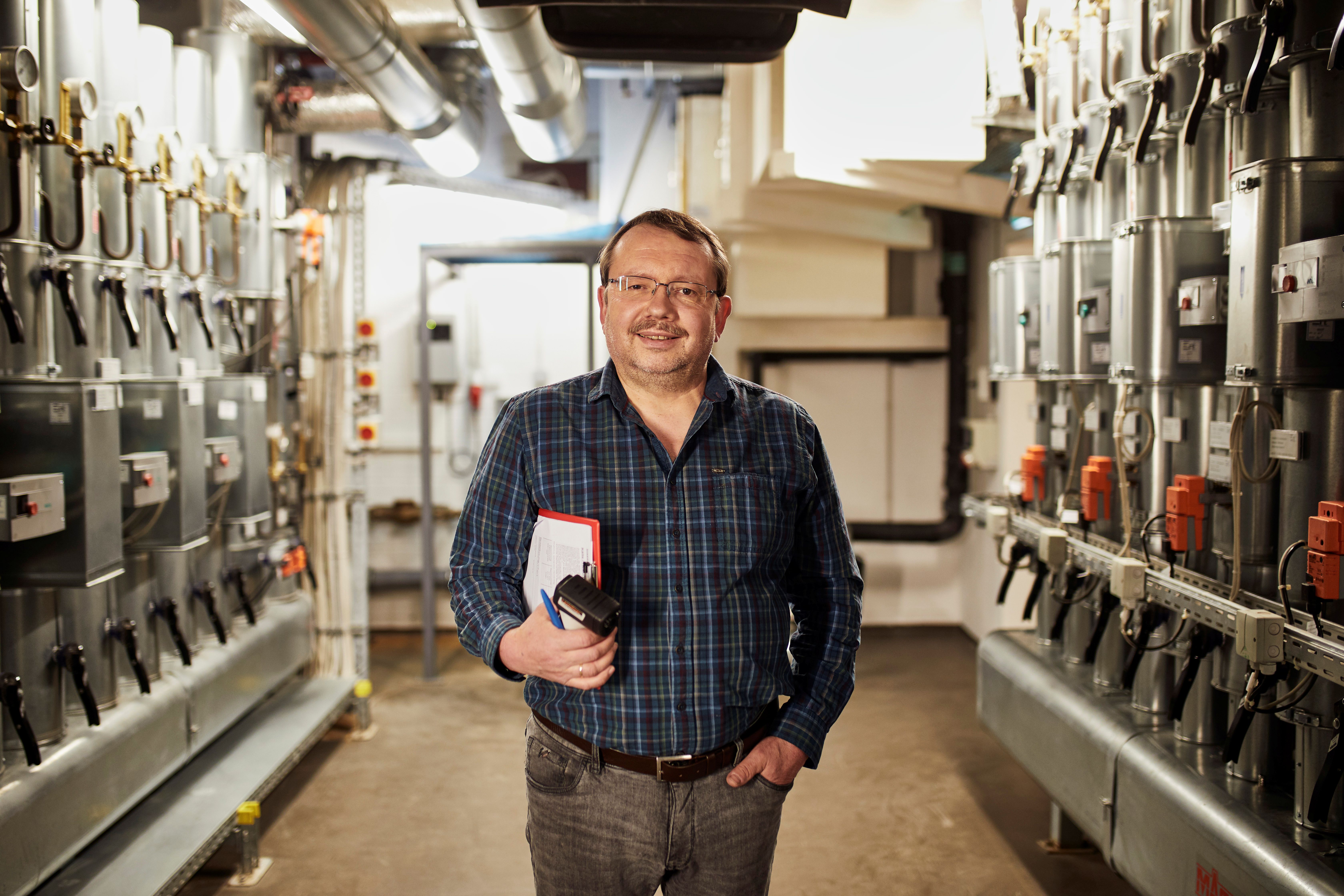 Herr Wruck steht in einem technischen Gang zwischen Industrieanlagen. Er hält ein Notizbuch.  Auf beiden Seiten sind Rohre, Ventile und Steuerungselemente zu sehen.