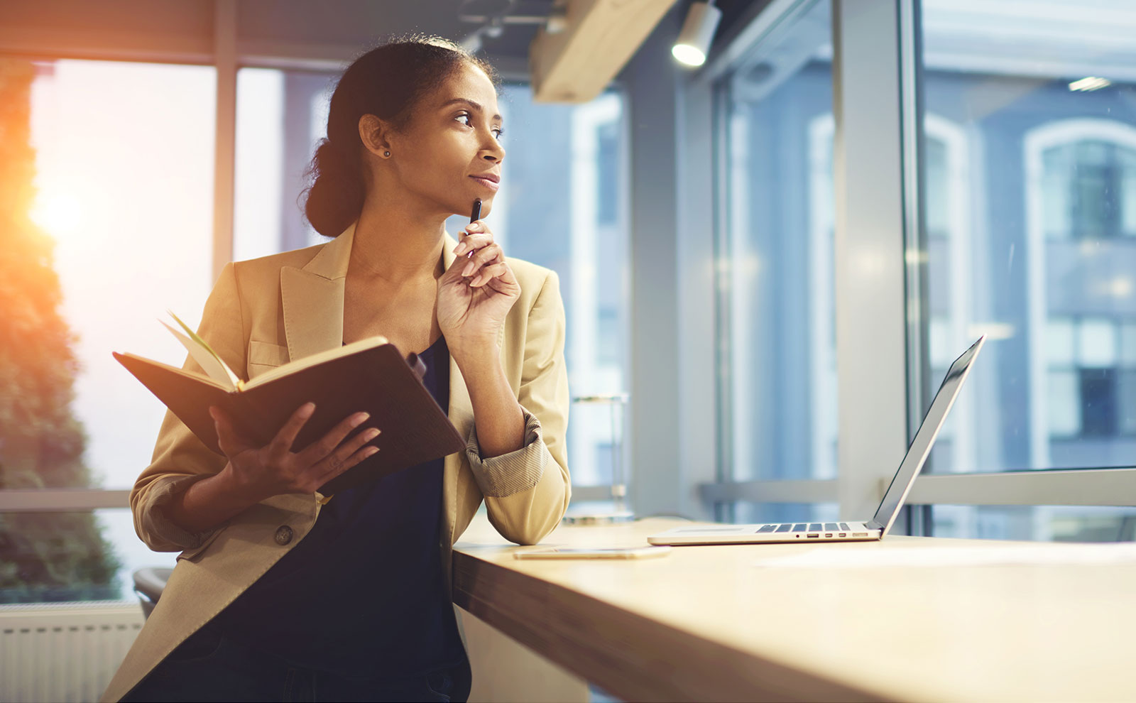 Weibliche Person stützt sich seitlich auf einen Tisch, auf dem ein Laptop steht. Sie strägt einen beigen Blazer, hat ein Notizbuch und Stift in der Hand und guckt nachdenklich nach links.