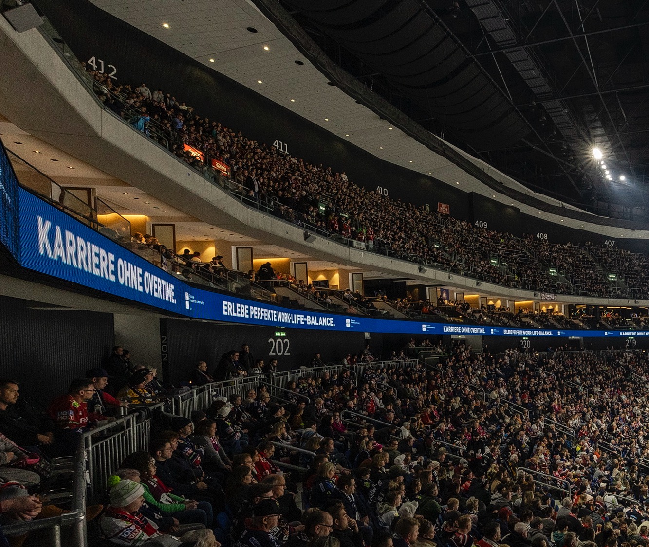 Mittlere Ränge der vollen Uber-Arena und VIP-Lounges. Auf dem Fascia-Board steht weiß auf blauem Hintergrund: Karriere ohne Overtime - Erlebe perfekte Work-Life-Balance.