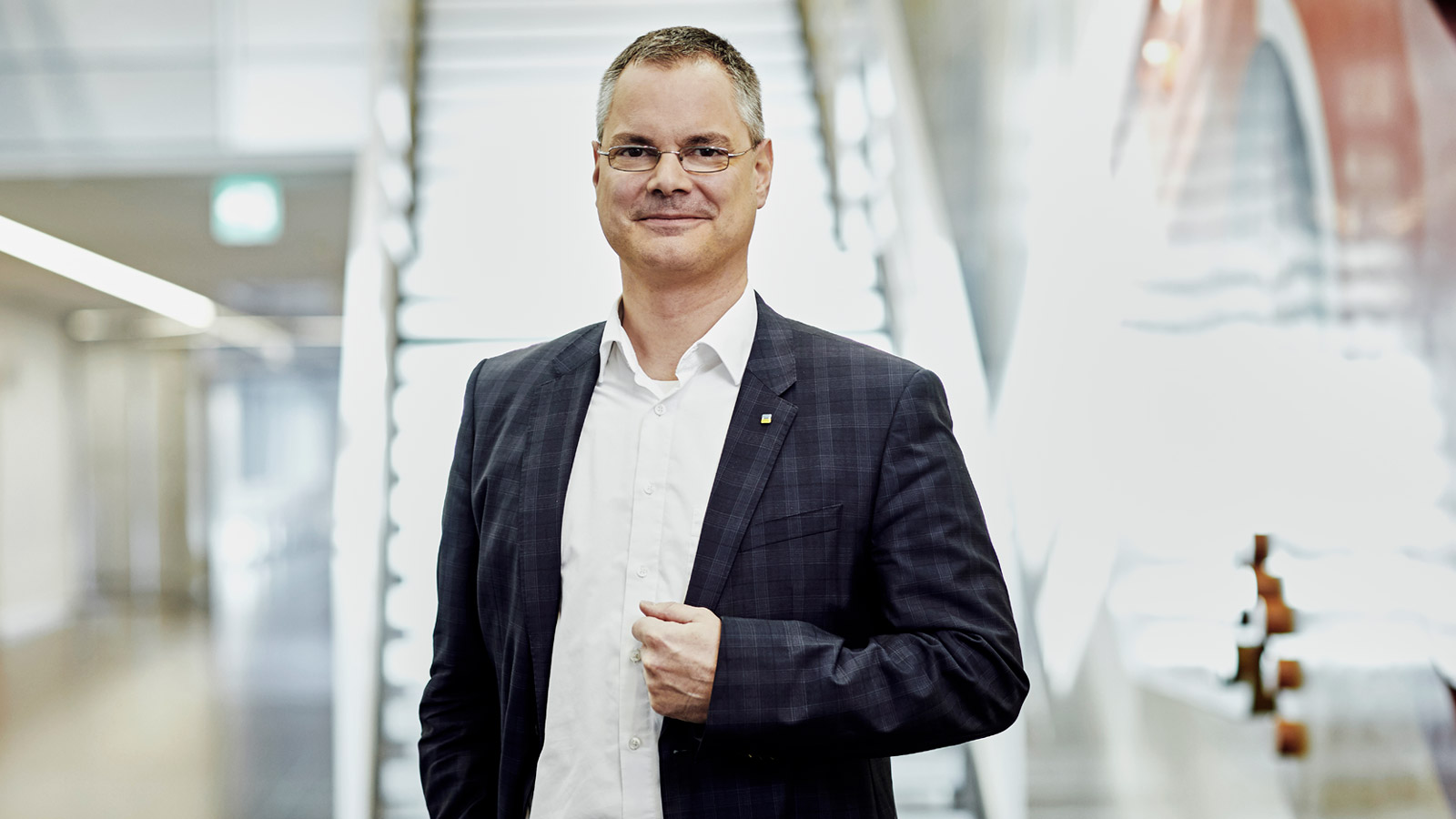 Ein freundlich guckender älterer Mann in einem schwarz-grau kariertem Anzug und Brille steht vor einer weißleuchtenden Treppe in einem Bürogebäude.