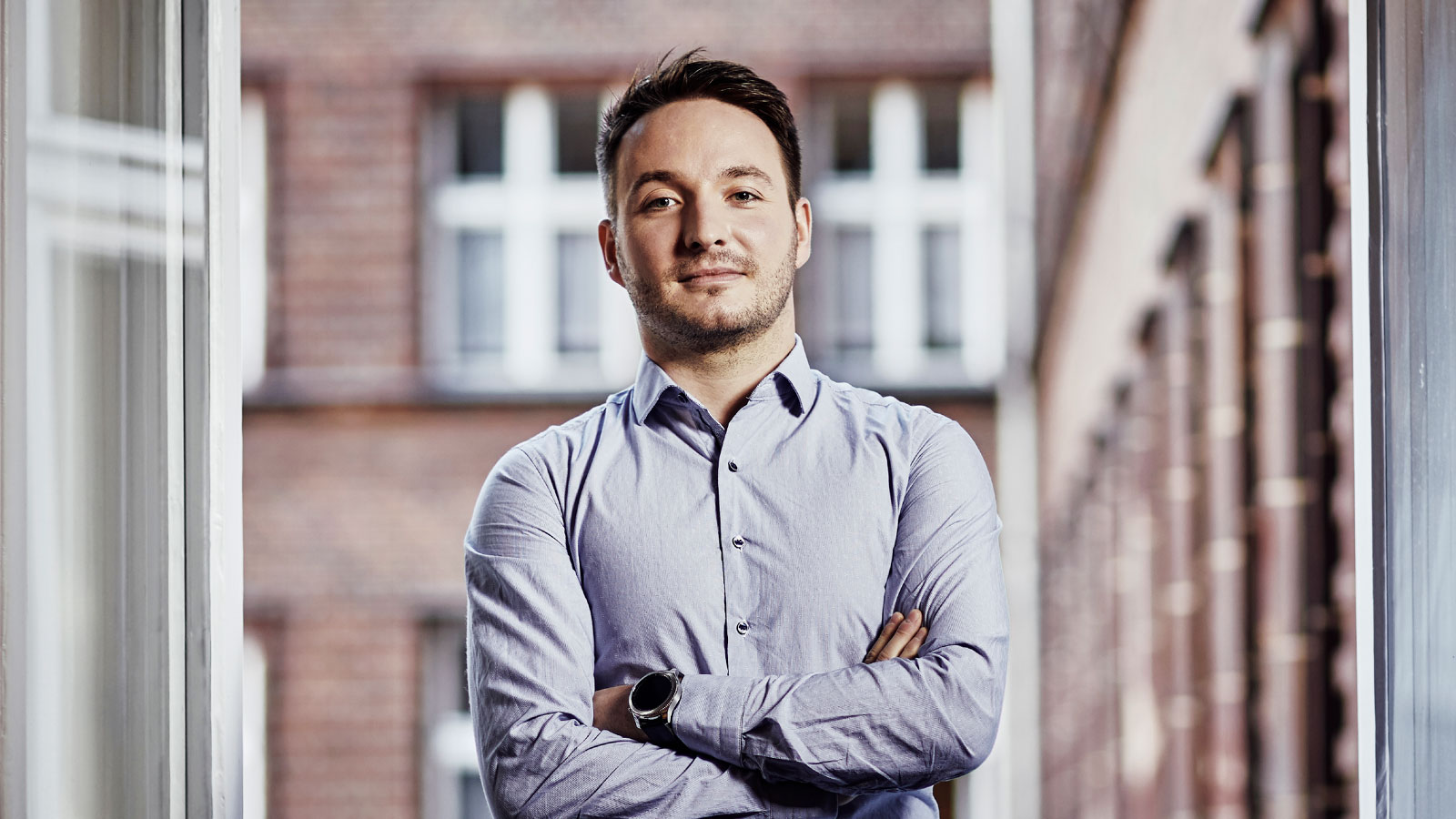 Ein Verwaltungsmitarbeitender guckt selbstbewusst und zufrieden in die Kamera. Er tägt ein hellblaues Hemd und steht vor einem Fenster.