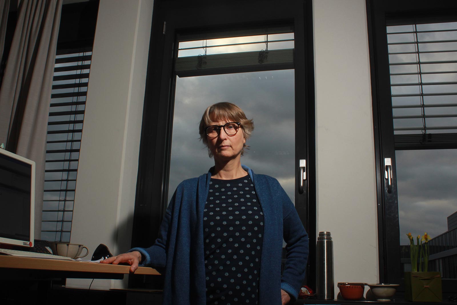 Eine weibliche Person steht in einem Bürogebäude vor Fenstern. Sie hat ihren Arm auf einem Schreibtisch, auf diesem sind eine Tasse und ein Monitor. Sie trägt eine blaue Strickjacke und hat eine Brille.