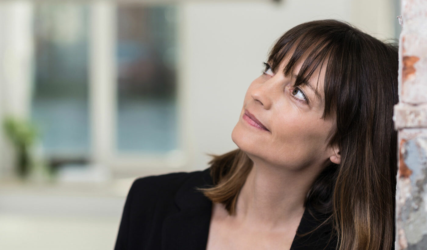 Weibliche Person mit braunen mittellangen Haaren und Pony guckt neugierig nach links oben. Sie trägt schwarze Kleidung, der Hintergrund ist verschwommen.