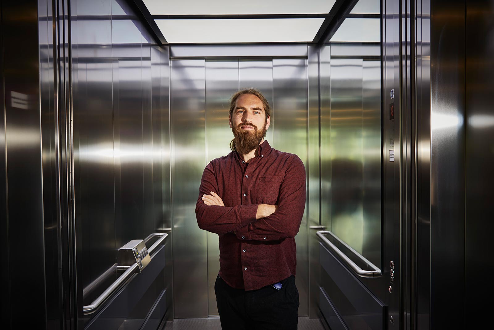 Männliche Person steht leicht lächelnd in einem silbernen Fahrstuhl. Er hat die Arme verschränkt, zurückgebundene Haare und einen Bart und trägt ein dunkerotes Hemd.