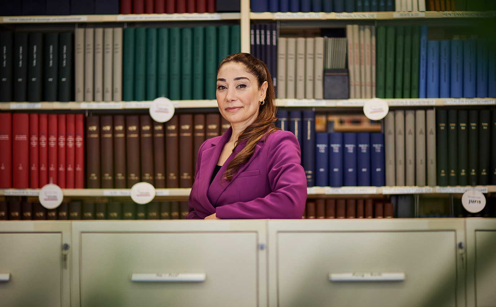 Frau mit braunen, zum Zopf gebundenen Haaren steht in einem Büro. Sie trägt einen lilanen Anzug, stützt sich auf Aktenschränke und steht vor einer Bücherwand.