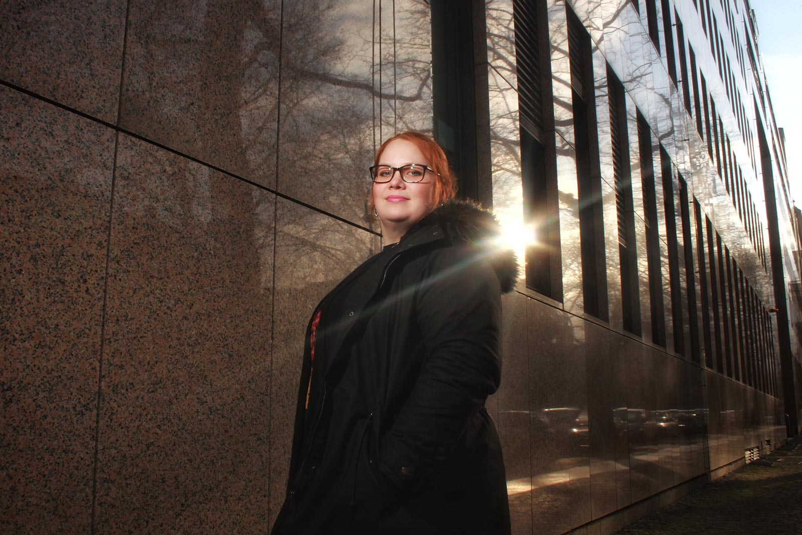 Eine weibliche Person mit roten, zurückgebundenen Haaren steht in einem schwarzen Mantel vor einer Gebäudefassade. Von den Fenstern spiegeln sich die Sonne, Bäume und Autos.