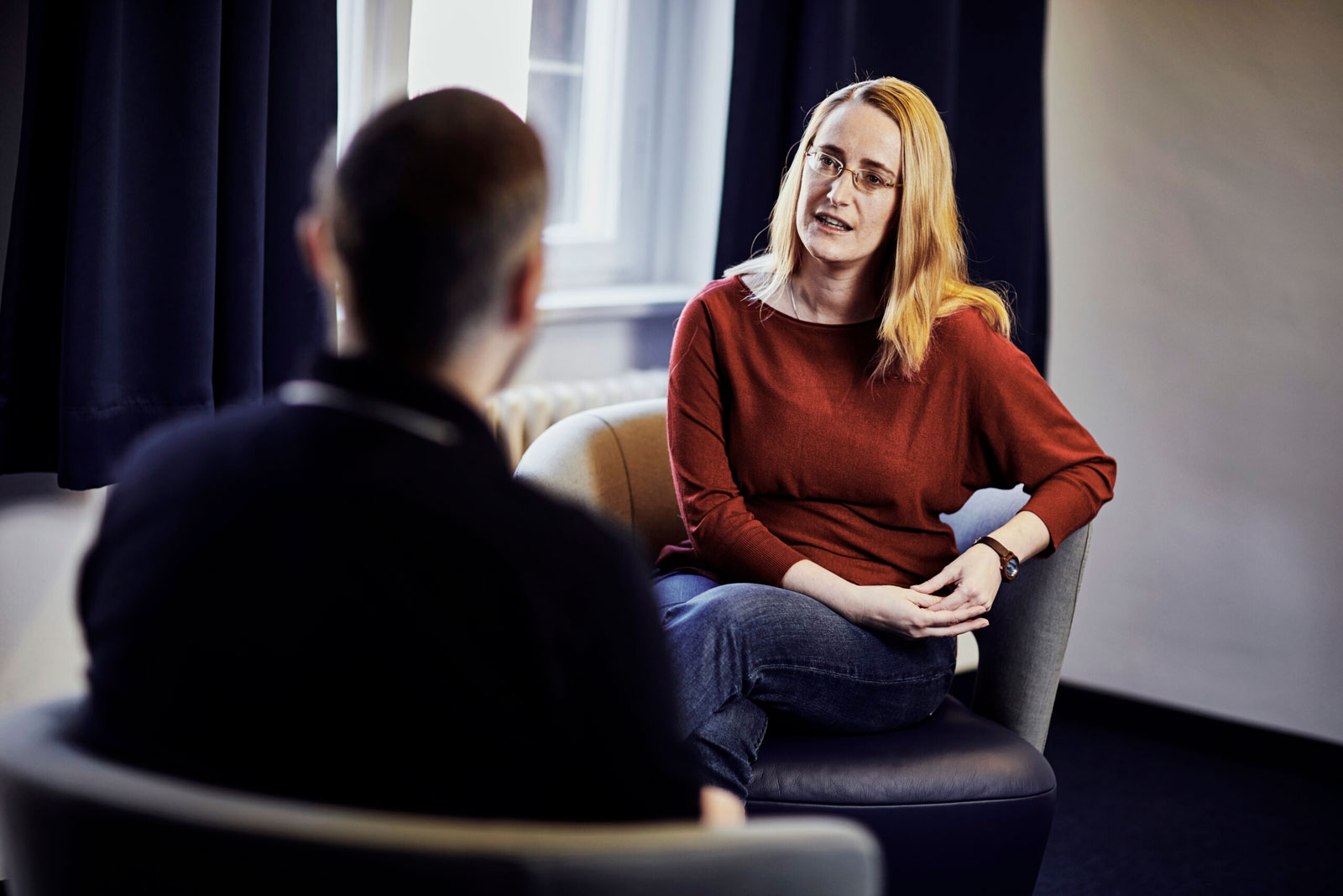 Zwei Personen sitzen einander in Sesseln gegenüber, eine mit dem Rücken zur Kamera, die andere mit dem Gesicht. Sie führen ein ernstes Gespräch.