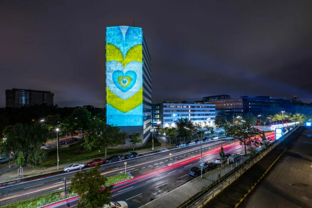 Ein hohes Gebäude bei Nacht mit einer beleuchteten Projektion in Herzform in Blau, Gelb und Weiß. Im Vordergrund sind Lichtspuren vorbeifahrender Autos auf einer Straße zu sehen.
