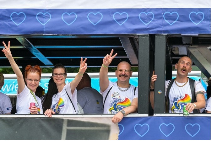 Vier lachende DRV Bund Mitarbeitende beim CSD 2025 stehen auf einem Truck vor einem Herzbanner. Sie tragen bunte Shirts und zeigen Peace-Zeichen.