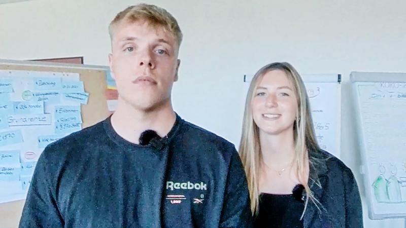 Zwei duale Studierende stehen in einem Raum mit Pinnwänden im Hintergrund. Sie tragen schwarze Kleidung.