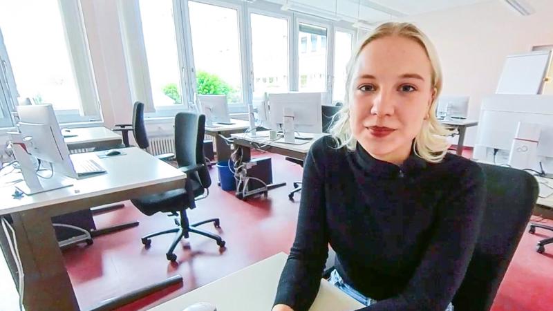 Duale Studentin sitzt in einem Klassenzimmer. Sie trägt einen schwarzen Pullover, im Hintergrund sind Schreibtische und Monitore zu sehen.