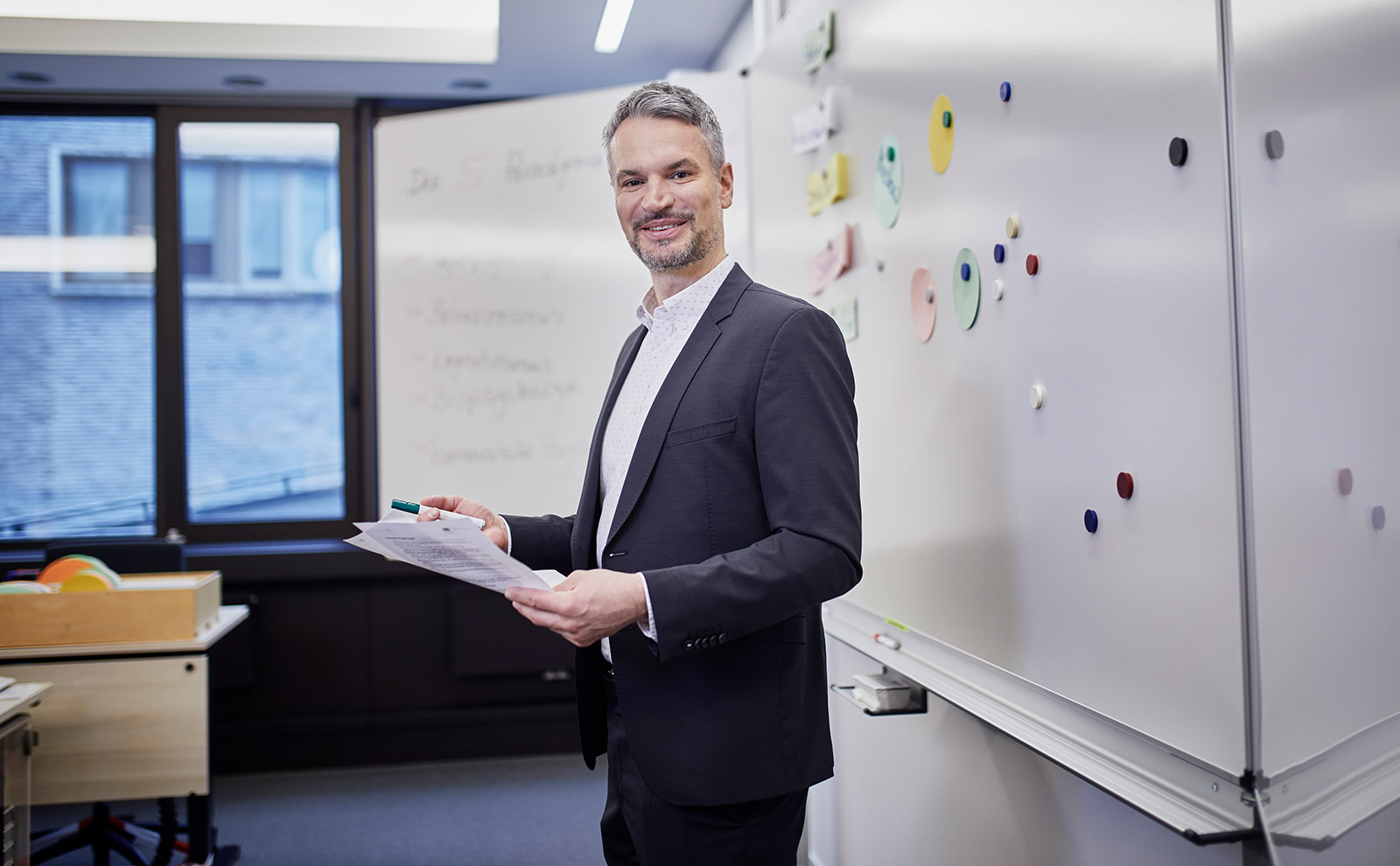 Prof. Dr. Breitling steht in einem dunklen Anzug vor einem Whiteboard in einem Klassenzimmer. Er hat Papier und Stift in der Hand und auf dem Whiteboard sind Zettel und Schrift zu erkennen.