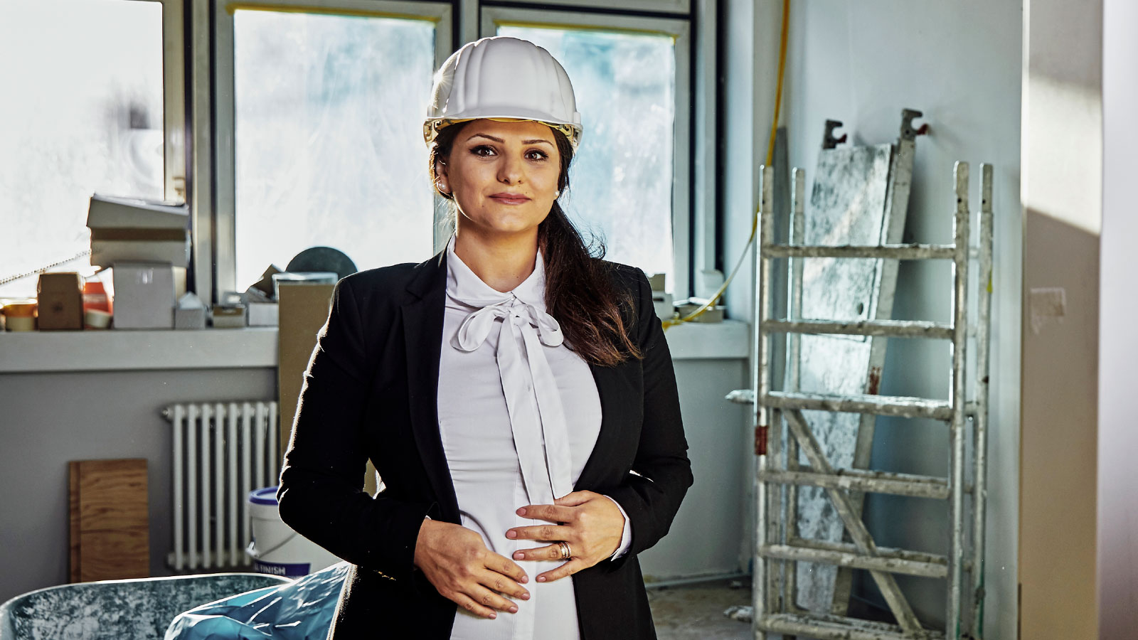 Lana steht in einem hellen Raum mit Baustellencharakter. Sie trägt einen weißen Schutzhelm, eine weiße Bluse und einen Blazer. Hinter ihr stehen Bauutensilien wie eine Leiter.