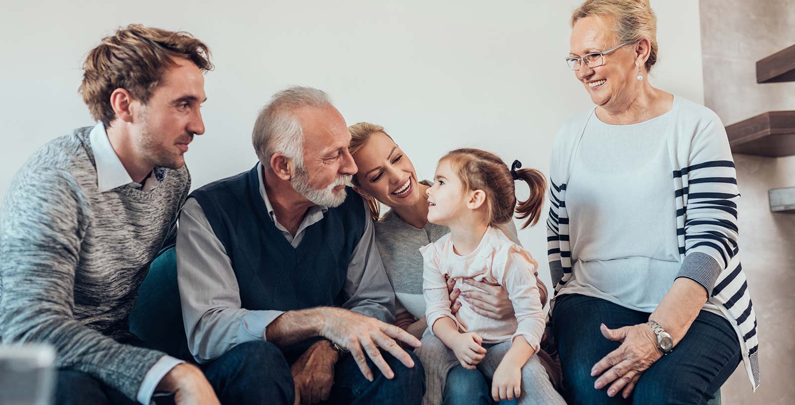Mehrgenerationenfamilie auf dem Sofa: Großeltern, Mutter und junger Mann lachen mit einem kleinen Mädchen im Wohnzimmer.
