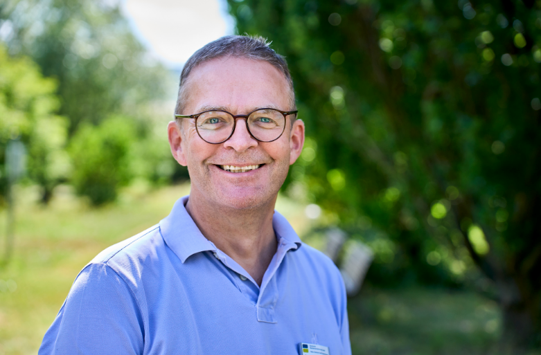 Ein älterer Arzt im weißen Kittel und mit Stethoskop steht lächelnd vor einer Klinikfassade und schaut in den Himmel.