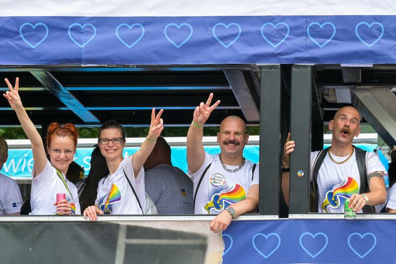 Gruppe von Personen mit Regenbogen-T-Shirts auf einem Balkon; zeigen Friedenszeichen, im Hintergrund ein blaues Banner mit Herzsymbolen.