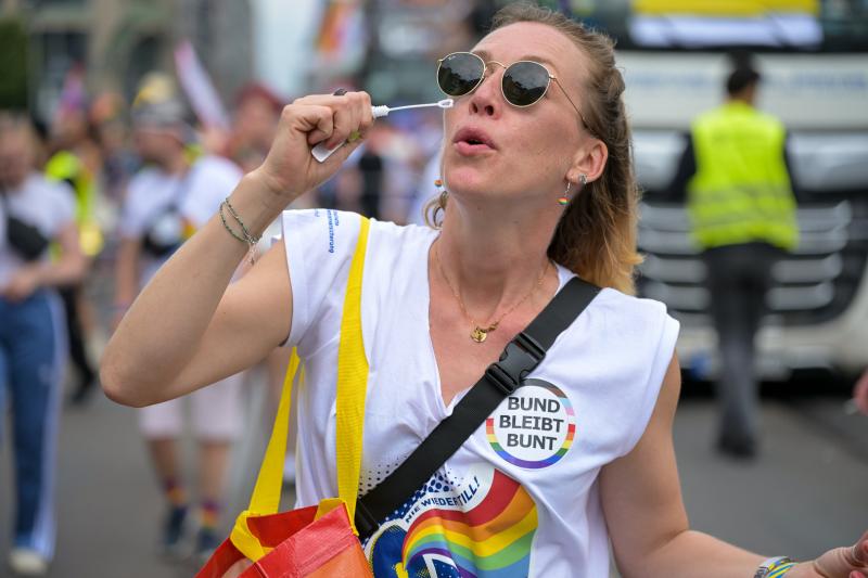 Person mit Regenbogen-Shirt und „BUND BLEIBT BUNT“-Badge bei öffentlicher Veranstaltung; hält Seifenblasenstab, im Hintergrund weitere Teilnehmende und ein Polizeifahrzeug