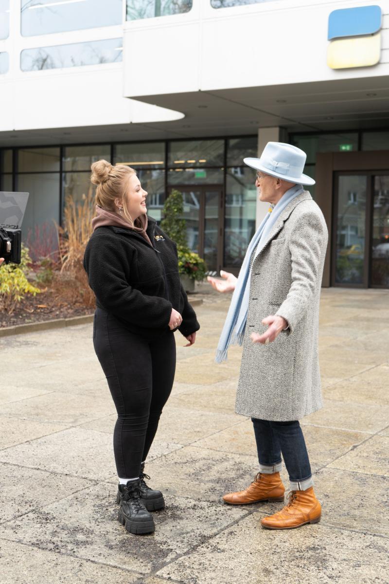 Zwei Personen (Günther Krabbenhöft und eine Mitarbeitende der DRV Bund) im Gespräch vor einem DRV Bund Gebäude; tragen wettergerechte Kleidung mit Jacken, Mützen und Schals.