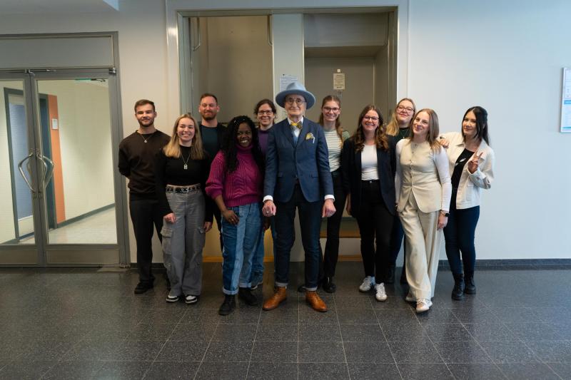 9 Mitarbeitende der DRV und Günther Krabbenhöft stehen in einem Flur vor Doppeltüren; einige stehen, andere hocken leicht, links Glasdurchgang sichtbar.