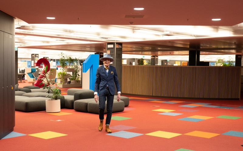 Günther Krabbenhöft im Anzug geht durch modernes Büro mit buntem Teppich, Sitzbereich, Pflanzen und großen Zahlen als Dekoration.