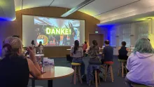 Mehrere Personen sitzen auf Hockern in einem Konferenzraum und blicken auf eine große Leinwand. Auf dem Bildschirm steht in gelben Buchstaben "DANKE!". Der Raum ist modern beleuchtet mit blauen und gelben Lichtakzenten.