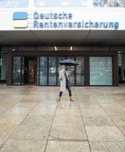 Ein älterer Mann steht tanzend mit einem Regenschirm vor einem Bürogebäude, auf welchem "Deutsche Rentenversicherung Bund" steht. Er trägt einen Jackett, Fliege, einen grauen Hut und einen langen grauen Mantel.