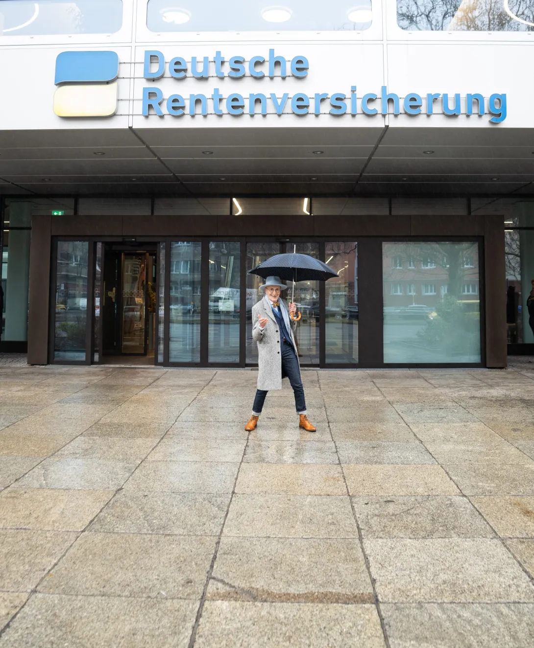 Ein älterer Mann steht tanzend mit einem Regenschirm vor einem Bürogebäude, auf welchem "Deutsche Rentenversicherung Bund" steht. Er trägt einen Jackett, Fliege, einen grauen Hut und einen langen grauen Mantel.